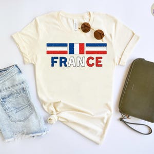 May include: Cream-colored t-shirt with the word "FRANCE" in blue and red block letters. Above the word is a graphic of the French flag. The shirt is paired with denim shorts, sunglasses, and a green clutch.