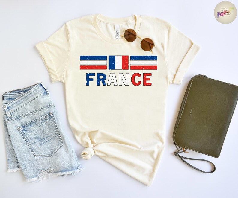 May include: Cream-colored t-shirt with the word "FRANCE" in blue and red block letters. Above the word is a graphic of the French flag. The shirt is paired with denim shorts, sunglasses, and a green clutch.