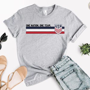 Może przedstawiać: Szara koszulka z napisem "ONE NATION. ONE TEAM." i emblematem USA. Projekt zawiera niebieskie, czerwone i białe paski. Koszulka jest połączona z dżinsowymi szortami, okularami przeciwsłonecznymi i sandałami.
