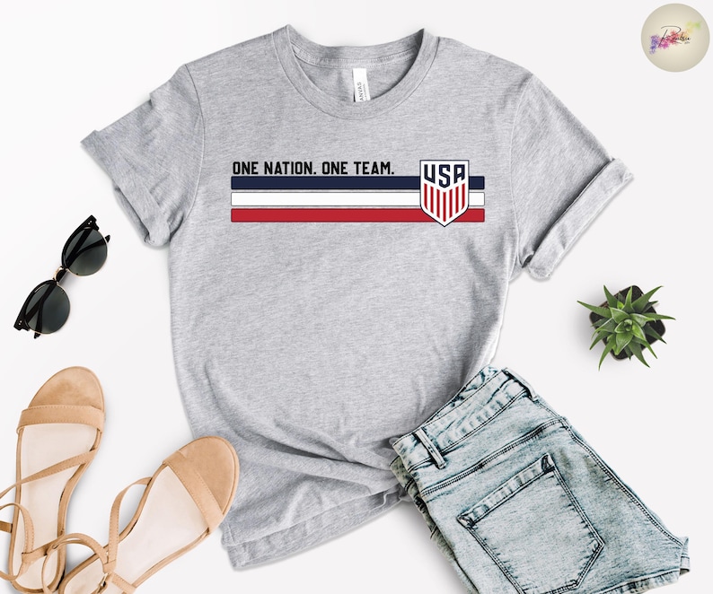Może przedstawiać: Szara koszulka z napisem "ONE NATION. ONE TEAM." i emblematem USA. Projekt zawiera niebieskie, czerwone i białe paski. Koszulka jest połączona z dżinsowymi szortami, okularami przeciwsłonecznymi i sandałami.