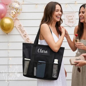 Könnte beinhalten: Eine schwarze Tragetasche mit dem Namen Clara in weißer Schreibschrift. Die Tasche hat zwei schwarze Schulterriemen und drei äußere Netztaschen, die eine Tube, ein Smartphone und eine weiße Flasche enthalten. Die Tasche besteht aus schwarzem Stoff.