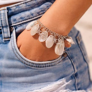 Könnte beinhalten: Ein silbernes Armband mit weißen Muschelanhängern und kleinen Seestern-Anhängern. Das Armband wird am Handgelenk getragen, die Hand ruht in der Tasche einer blauen Jeansshorts.