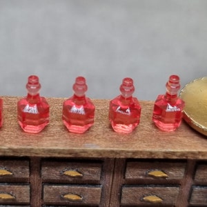 May include: Six miniature glass bottles containing a clear, red liquid. The bottles have a rounded, octagonal shape and a small, round stopper. The bottles are arranged in a row on a wooden surface.