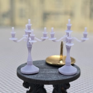 Könnte beinhalten: Miniatur-Kandelaber und eine kleine goldfarbene Schale stehen auf einem dunkelgrauen Tisch mit verzierten Beinen. Die Kandelaber sind helllila und haben mehrere Arme für Kerzen. Der Tisch steht auf einer karierten Oberfläche.