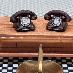 May include: Two miniature black rotary dial phones with silver accents are displayed on a wooden shelf. The phones are identical and have a vintage look.