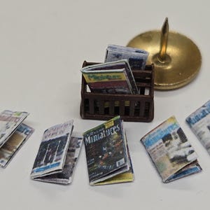 Può includere: Un set di riviste in miniatura con copertine colorate recanti il testo Miniatures, disposte attorno a un piccolo portariviste in plastica marrone. Una singola puntina da disegno in metallo color ottone è posizionata accanto al supporto.