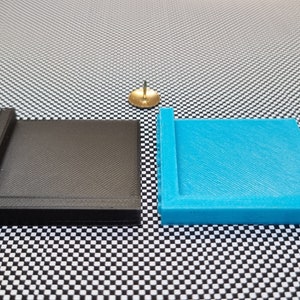 May include: Two 3D printed square trays, one black and one blue, with a raised edge around the perimeter. The trays are on a black and white chequered surface.
