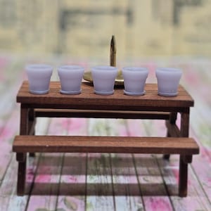 Op de afbeelding: Miniatuur houten picknicktafel met een bank, met vijf kleine, lavendelkleurige potten. Een messing kandelaar met een enkele kaars staat achter de potten. De achtergrond heeft een bloemen- en patroonontwerp.