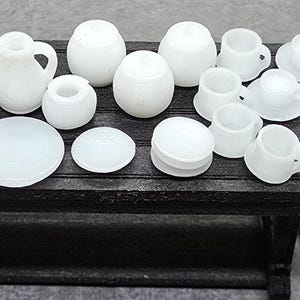 May include: A miniature white porcelain tea set, including a teapot, creamer, sugar bowl, plates, cups, and saucers, arranged on a small, dark wooden table. The set is displayed against a neutral grey background, showcasing the delicate details of each piece.