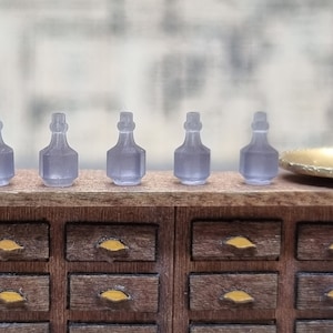Könnte beinhalten: Fünf durchsichtige Glasflaschen mit runder, bauchiger Form und schmalem Hals, in einer Reihe auf einem Holzregal mit Schubladen angeordnet. Die Flaschen sind helllila und haben eine matte Oberfläche.