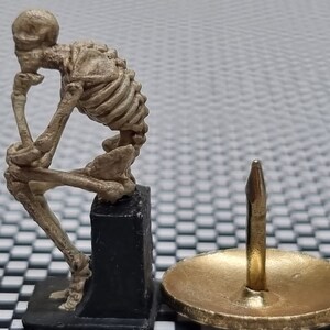 Könnte beinhalten: Eine kleine, beige Skelettfigur, die auf einem schwarzen Sockel sitzt, mit einer goldenen Stecknadel auf einer goldenen Basis im Hintergrund.