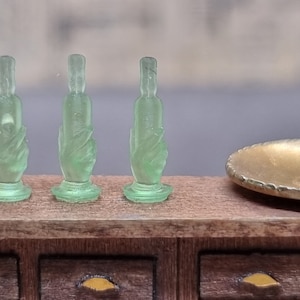 May include: Three clear green glass bottle stoppers with a hand design. The stoppers are sitting on a wooden surface.