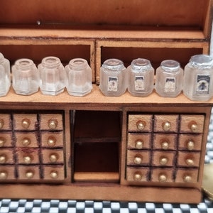 May include: A miniature wooden apothecary cabinet with drawers and a shelf holding several small, clear glass jars. The jars have various shapes and some have labels. The cabinet has gold-coloured knobs on the drawers.