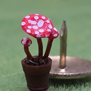 May include: A miniature brown clay pot containing three small red mushrooms with white spots on their caps. The mushrooms have thin brown stems and are planted in dark soil. A metal push pin stands upright next to the tiny potted plant.