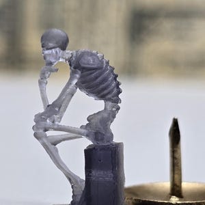 May include: A clear plastic figurine of a skeleton sitting in a thinking pose on a small base. The figurine is a replica of the famous "The Thinker" statue.