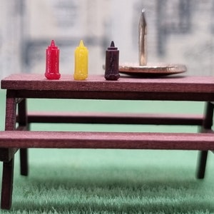 Puede incluir: Pequeña mesa de picnic de madera con tres botellas de condimentos en miniatura: ketchup rojo, mostaza amarilla y salsa barbacoa marrón. Una chincheta grande está sobre la mesa. La mesa es de madera oscura y está sobre una superficie verde.
