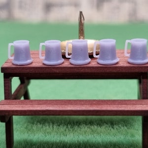 May include: Five miniature lavender-colored beer mugs with handles are arranged on a small, dark brown picnic table. The table has a matching bench. A gold bottle cap and a silver tack are behind the mugs. The background is green.
