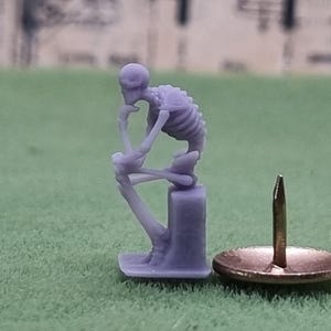 May include: A small, purple, 3D printed figurine of a skeleton sitting in a thinking pose. The figurine is on a small, purple base. A small, silver push pin is next to the figurine.