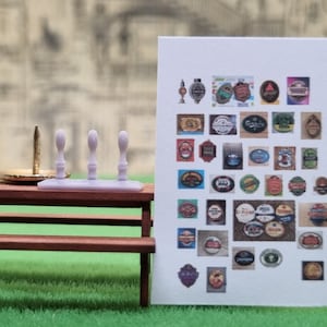 May include: A set of four miniature pub taps, three are white and one is gold, on a wooden table. A sheet of paper with a grid of miniature beer labels is in the background.