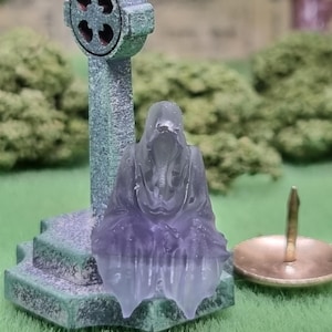 Może przedstawiać: Miniaturowa scena cmentarna z zwietrzałym, zielono-szarym nagrobkiem w kształcie krzyża i przezroczystą, fioletowo-przezroczystą postacią ducha. W pobliżu na trawiastym podłożu znajduje się mały, mosiężny świecznik. Scena przywodzi na myśl upiorny, gotycki motyw.