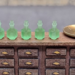 May include: Five small, clear green glass bottles with a rounded, bulbous shape and a hexagonal base. The bottles are arranged in a row on a wooden surface.