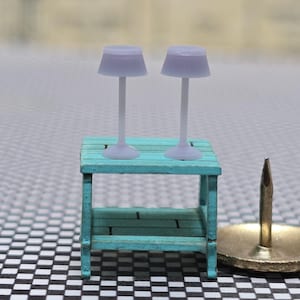 May include: Two miniature lavender lamps with white stems are on a small, turquoise wooden table. A gold thumbtack is next to the table. The table has a lower shelf. The background is blurred.