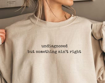 May include: A beige sweatshirt with the text "undiagnosed but something ain't right" printed on the front. The sweatshirt is a relaxed fit and has a slightly oversized look. The color of the sweatshirt is "SAND".