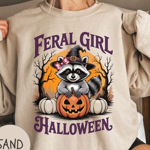 May include: Sand-coloured sweatshirt with a Halloween graphic. The design features a raccoon wearing a witch hat and holding a carved pumpkin. The text "FERAL GIRL" and "HALLOWEEN" are printed above and below the raccoon illustration.
