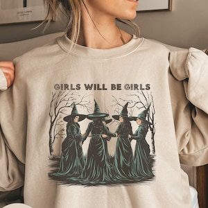 May include: A sand-coloured sweatshirt featuring a graphic of four witches in a circle, wearing black pointed hats and long green robes. The text "GIRLS WILL BE GIRLS" is printed above the witches.