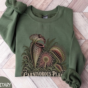 May include: A military green crewneck sweatshirt featuring a detailed graphic print of various carnivorous plants, including a pitcher plant and a Venus flytrap, with the text "CARNIVOROUS PLANTS" printed in a decorative font at the bottom.