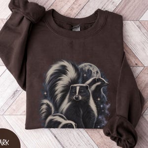 May include: A dark chocolate brown crewneck sweatshirt featuring a graphic print of three skunks against a starry night sky with a full moon. The long-sleeved pullover has a ribbed collar, cuffs, and hem, showcasing a detailed illustration.