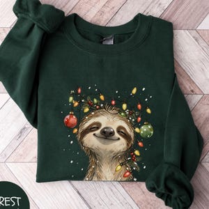 May include: A forest green long-sleeve sweatshirt featuring a graphic illustration of a smiling sloth tangled in colourful Christmas lights and ornaments. The design includes red and green baubles, glowing light bulbs, and white snowflakes.