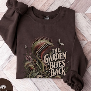 May include: A dark chocolate brown crewneck sweatshirt featuring a graphic print of a Venus flytrap plant with green and red leaves, a small insect, and the text "THE GARDEN BITES BACK" in a stylized, weathered cream-colored font on the front.
