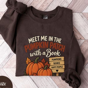 May include: A dark chocolate brown crewneck sweatshirt featuring a graphic with the text "MEET ME IN THE PUMPKIN PATCH with a Book" above two orange pumpkins and a wooden sign listing "PUMPKINS", "BOOKS", and "HAPPY PEOPLE" with a heart icon.