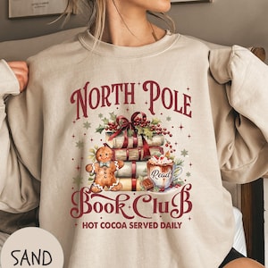 May include: Sand-coloured sweatshirt featuring a festive "North Pole Book Club" design. The graphic includes a stack of books, gingerbread man, and a cup of hot cocoa. The text "Hot Cocoa Served Daily" is also featured.