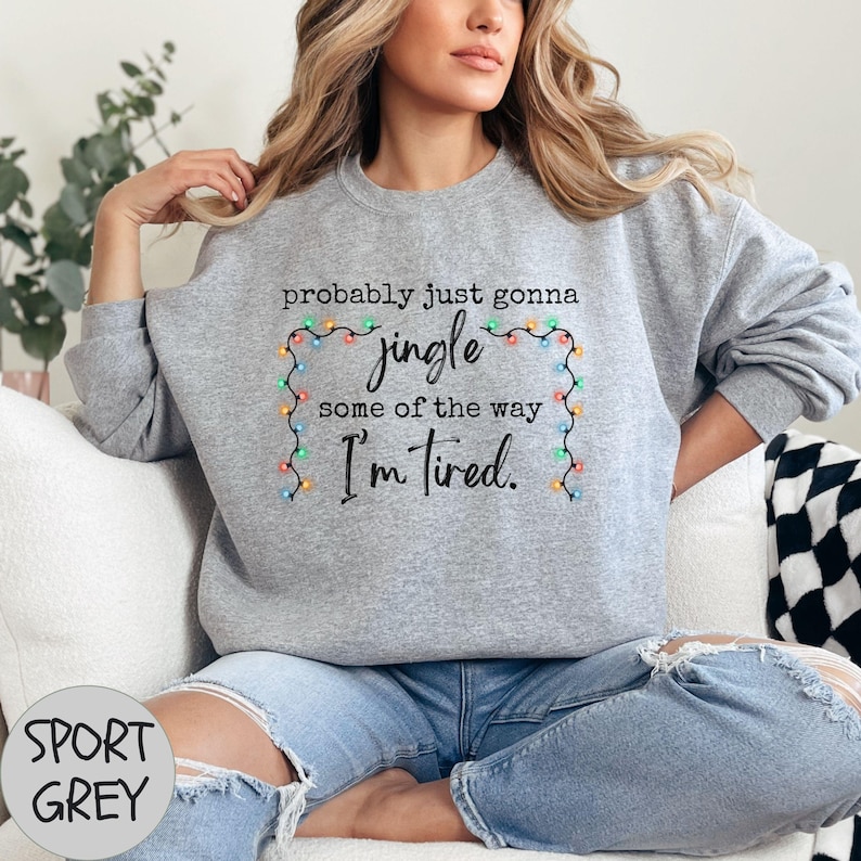 May include: Sport grey sweatshirt with the text "probably just gonna jingle some of the way I'm tired." The text is surrounded by a string of colorful Christmas lights. The sweatshirt is worn with blue jeans.