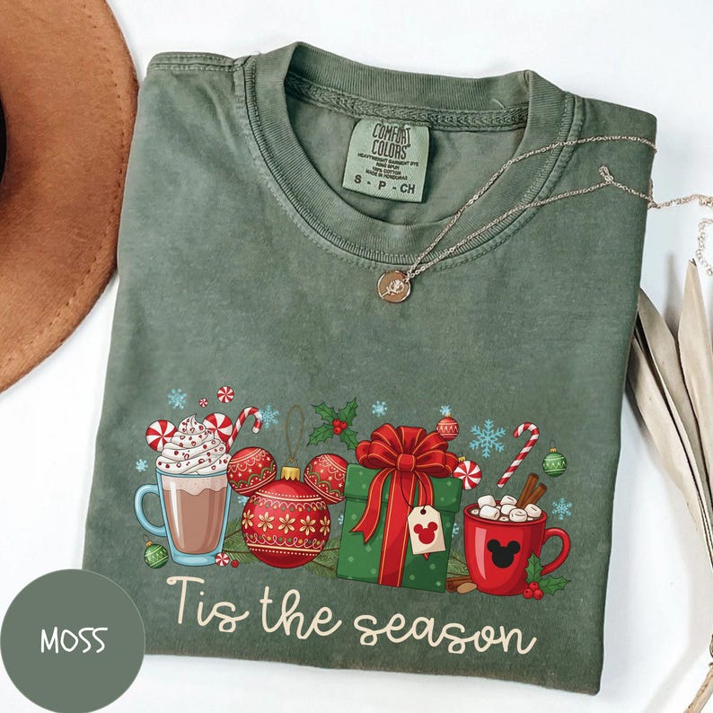 May include: Moss green Comfort Colors t-shirt with a festive graphic. The design includes hot cocoa, ornaments, a wrapped gift, and candy canes. The text "Tis the season" is written in a script font. A silver necklace is draped across the shirt.