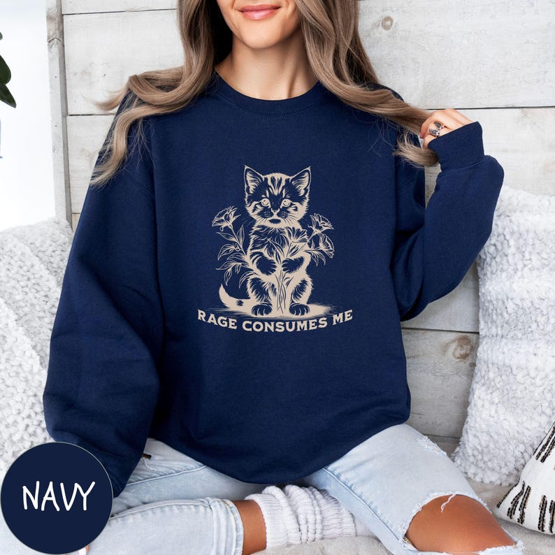 Puede incluir: Una sudadera azul marino con un gr&aacute;fico de un gato rodeado de flores. El texto "Rage Consumes Me" est&aacute; impreso debajo del gato.