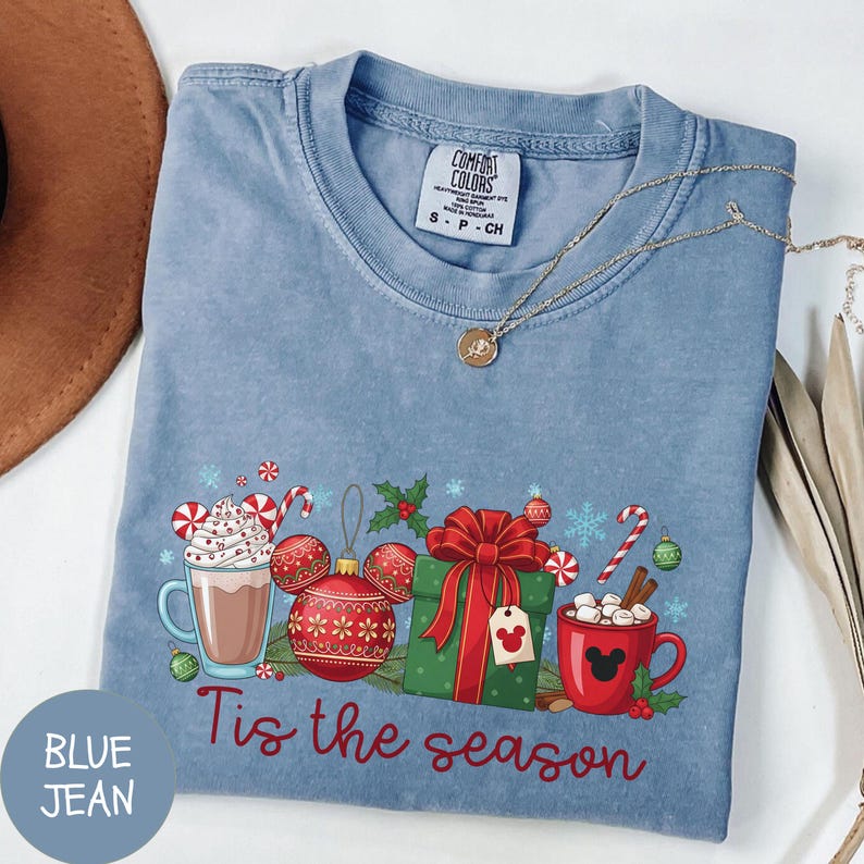 May include: Blue jean colored t-shirt with a festive graphic. The design includes a hot chocolate mug, Christmas ornaments, a wrapped gift, and the phrase "Tis the season" in red script. A gold necklace is draped across the shirt.