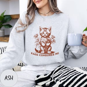 Puede incluir: Una sudadera gris con un gr&aacute;fico de un gato sosteniendo flores y el texto "Rage Consumes Me". La sudadera la lleva puesta una persona.