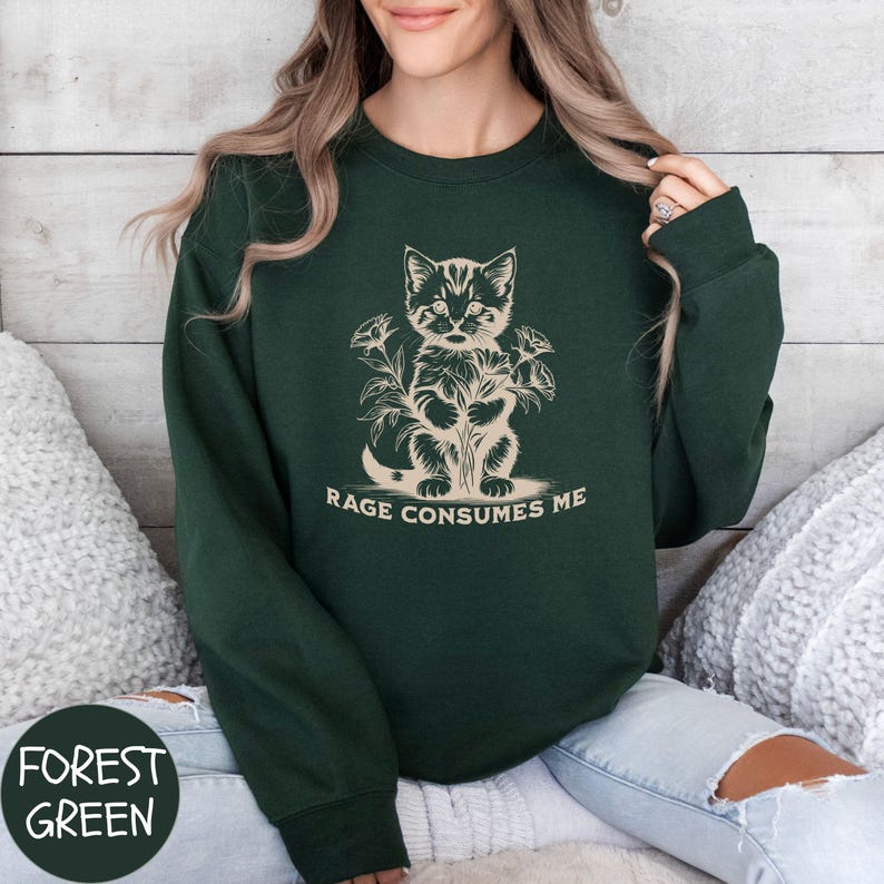 Puede incluir: Sudadera verde bosque con una ilustraci&oacute;n en blanco y negro de un gato con flores y el texto "Rage Consumes Me".