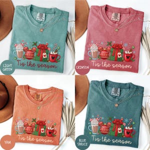 May include: Four different colored t-shirts with a holiday design. Each shirt features a graphic with a mug of hot chocolate, a Mickey Mouse ornament, a wrapped gift, and the text "Tis the season". The colors are light green, crimson, yam, and blue spruce.