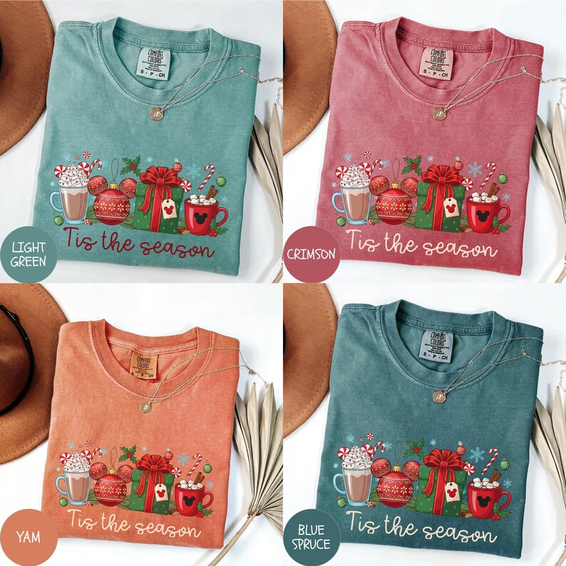 May include: Four different colored t-shirts with a holiday design. Each shirt features a graphic with a mug of hot chocolate, a Mickey Mouse ornament, a wrapped gift, and the text "Tis the season". The colors are light green, crimson, yam, and blue spruce.