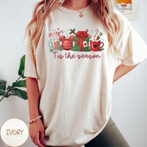 May include: Ivory-colored t-shirt featuring a festive design with hot cocoa, a Mickey Mouse ornament, a wrapped gift, and candy canes. The phrase "Tis the season" is printed in red script. The shirt is a relaxed fit.