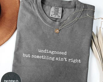 Puede incluir: Camiseta gris con el texto "undiagnosed but something ain't right" impreso en blanco.