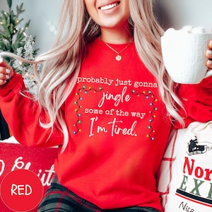 May include: A red long-sleeve sweatshirt with the text "probably just gonna jingle some of the way I'm tired." The text is surrounded by a string of colorful Christmas lights. A white mug is held in the hand.