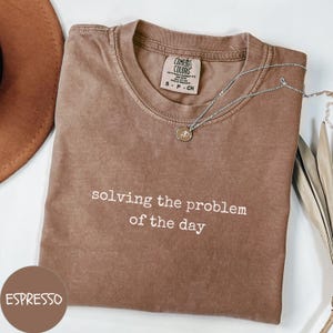 May include: A brown t-shirt with the text "solving the problem of the day" printed in white. The shirt is made of a soft, comfortable material and has a relaxed fit.