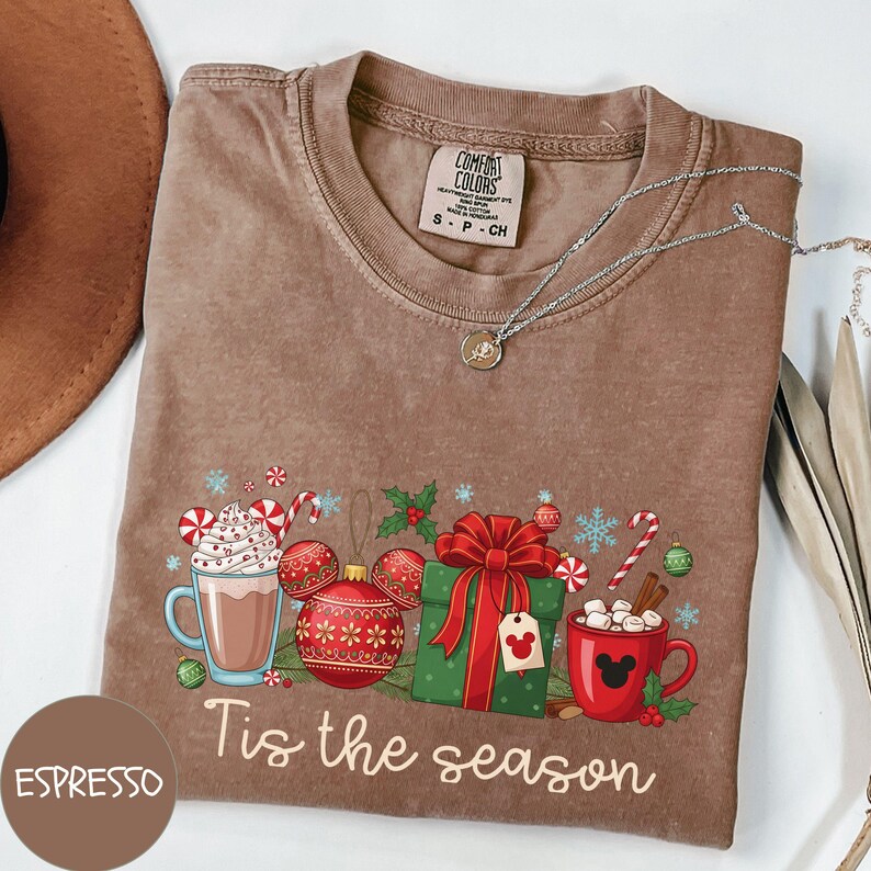 May include: Espresso-colored t-shirt with a festive graphic. The design includes a mug of hot chocolate, ornaments, a wrapped gift, and the text "Tis the season." A silver necklace is draped across the shirt.