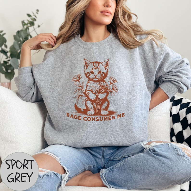 Puede incluir: Una sudadera gris con una ilustraci&oacute;n marr&oacute;n de un gato sosteniendo flores. El texto "RAGE CONSUMES ME" est&aacute; impreso debajo del gato.