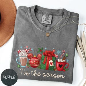 May include: A gray Comfort Colors t-shirt with a festive graphic. The design includes hot cocoa, ornaments, a wrapped gift, and candy canes. The text "Tis the season" is printed below the graphic. A silver necklace is draped across the shirt.